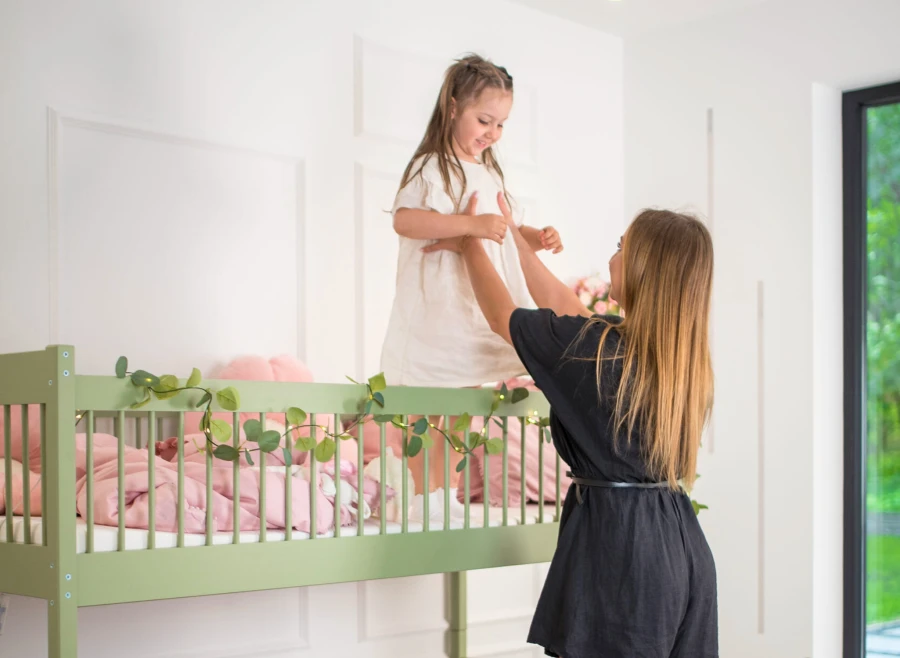 Mama wyjmująca uśmiechniętą dziewczynkę z oliwkowego łóżka piętrowego z barierkami.