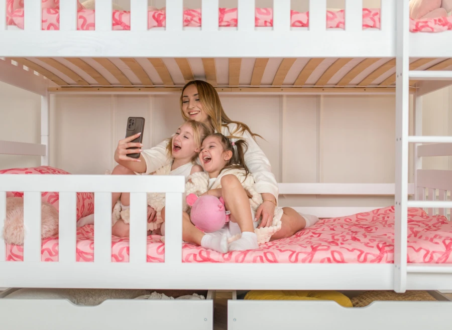 Mama i córki robią selfie na białym łóżku piętrowym z różową pościelą.
