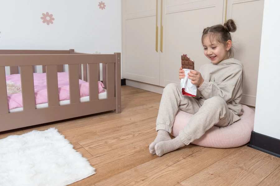 Uśmiechnięta dziewczynka siedzi na różowej pufie na podłodze, trzymając nadgryzioną tabliczkę czekolady. Obok widoczne niskie łóżko w kolorze mlecznej czekolady z różową pościelą. W tle beżowa szafa.