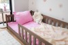 Niskie, drewniane łóżko dziecięce z barierką ochronną w kolorze mlecznej czekolady. Na łóżku leży różowa pościel i jasny, ażurowy kocyk, a obok barierki stoi poduszka w kształcie białego kwiatu.